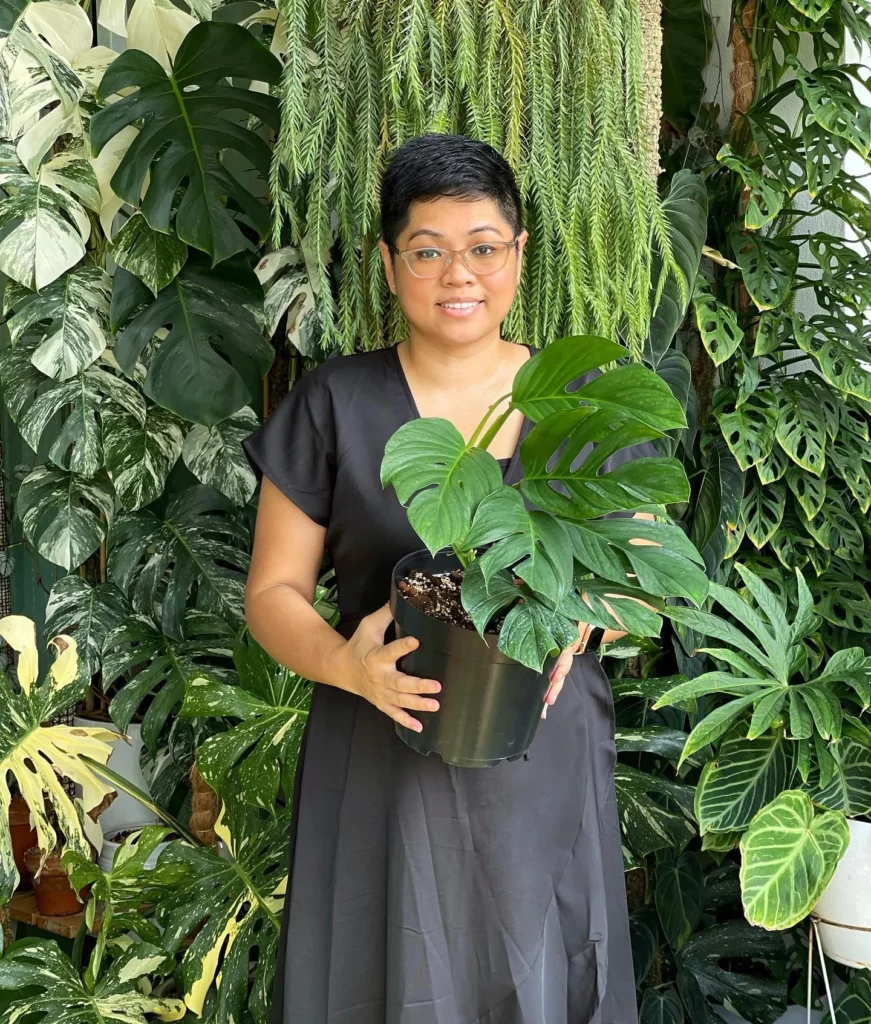 Happy Girl Posing with Monstera Pinnatipartita In 8" Black Pot with Other Indoor Plants Around Her In Garden