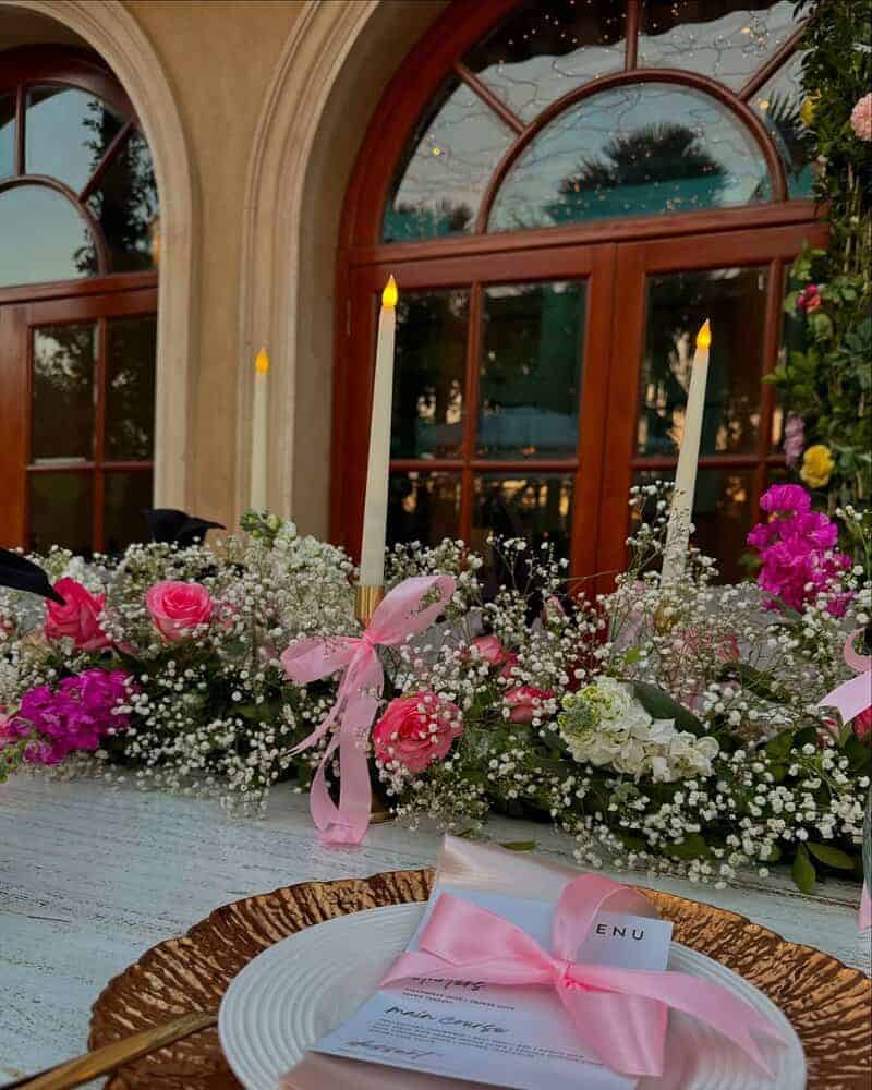 Outdoor table in front of arched wooden doors with a lush floral garland of pink flowers and baby’s breath, two tall white taper candles lit, and a place setting with a pink ribbon tied around a menu on a plate