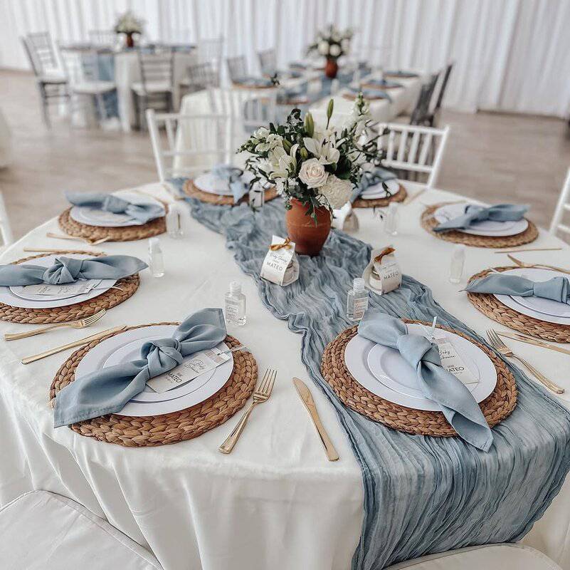 Round table with white linen and a dusty blue gauze runner, woven rattan chargers, white plates, dusty blue napkins tied in bows, gold flatware, small favor cartons, and a central vase of white roses and lilies