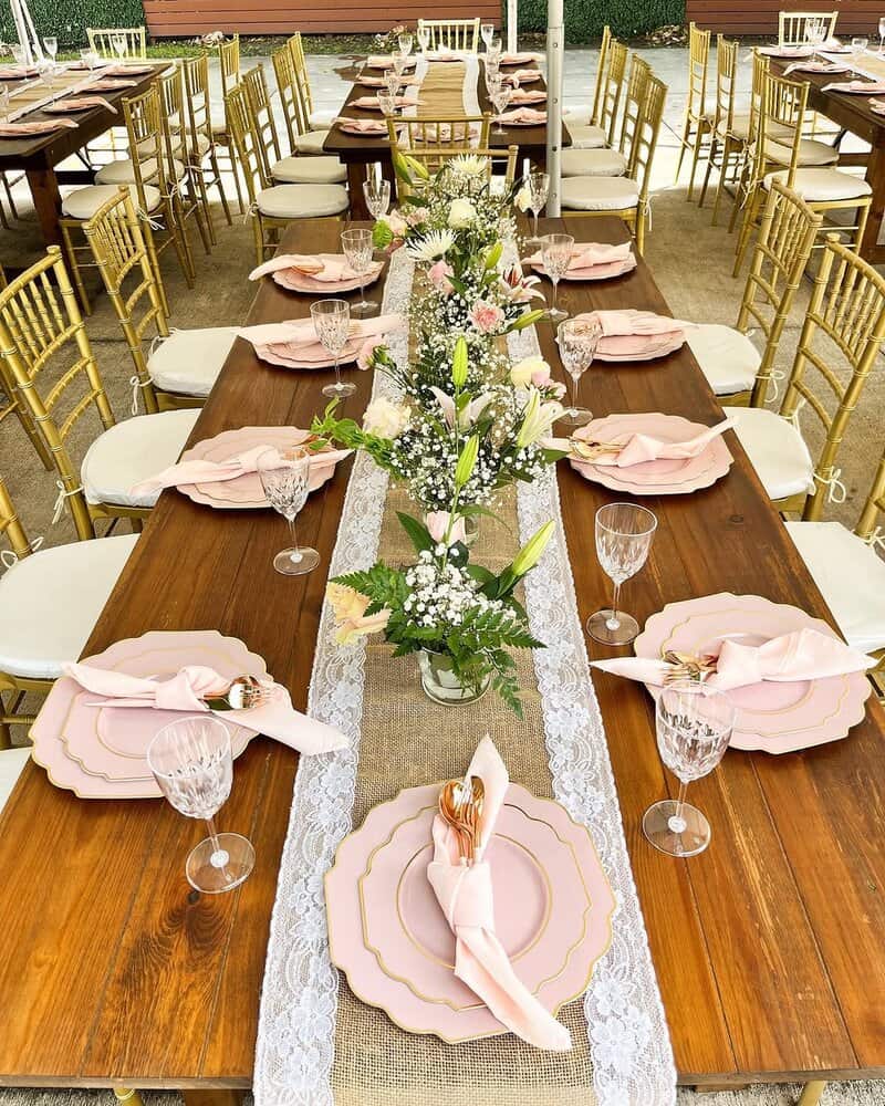 Long wooden farm tables under a tent with gold chiavari chairs, lace and burlap-style runner, clear goblets, pink scalloped plates with gold trim, blush napkins, and tall floral centerpieces in glass vases