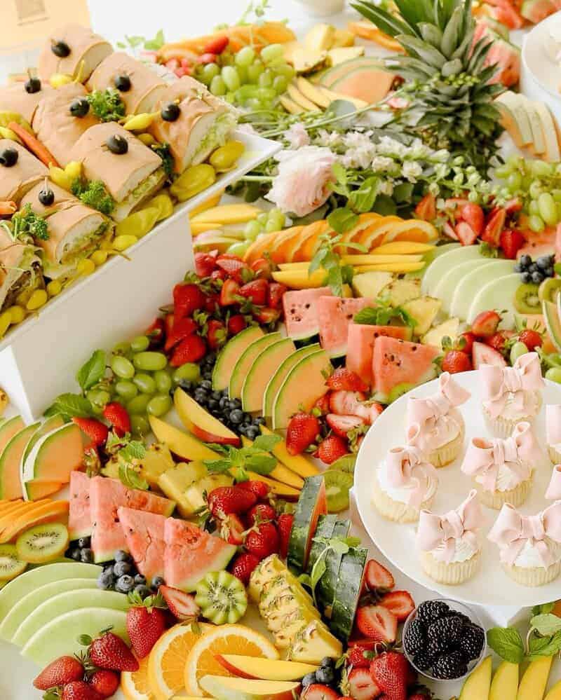 Overhead view of a bright grazing table with sliced watermelon, kiwi, grapes, strawberries, blueberries, mango, melon, pineapple, sandwiches on a white stand, and cupcakes topped with pale pink bows