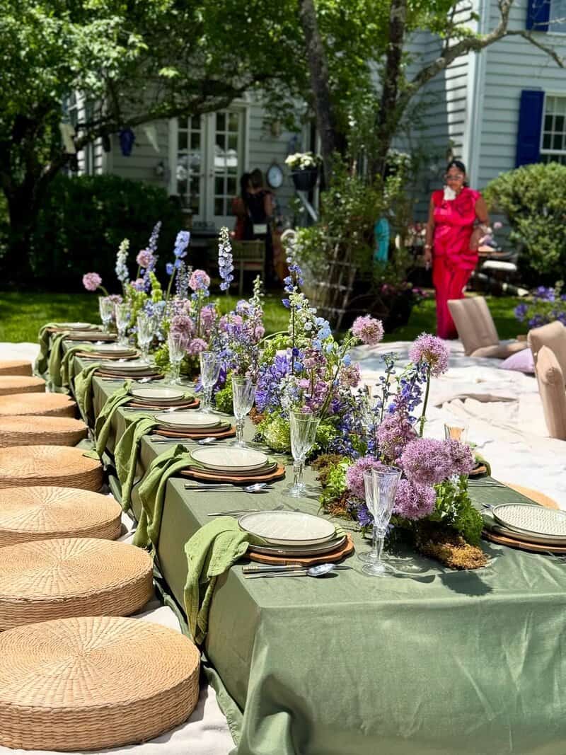 Outdoor long table with olive-green tablecloth and runner, woven round seat cushions, green plates, and a long arrangement of purple and blue flowers in clear glass vases under trees near a house