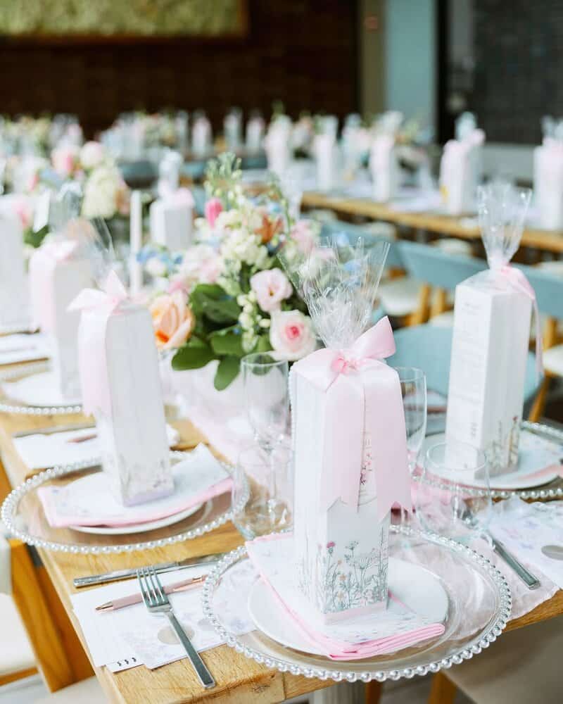 Long wooden tables set with beaded silver chargers, pink napkins, clear glassware, floral centerpieces, and tall rectangular favor boxes wrapped with pale pink ribbons at each place setting