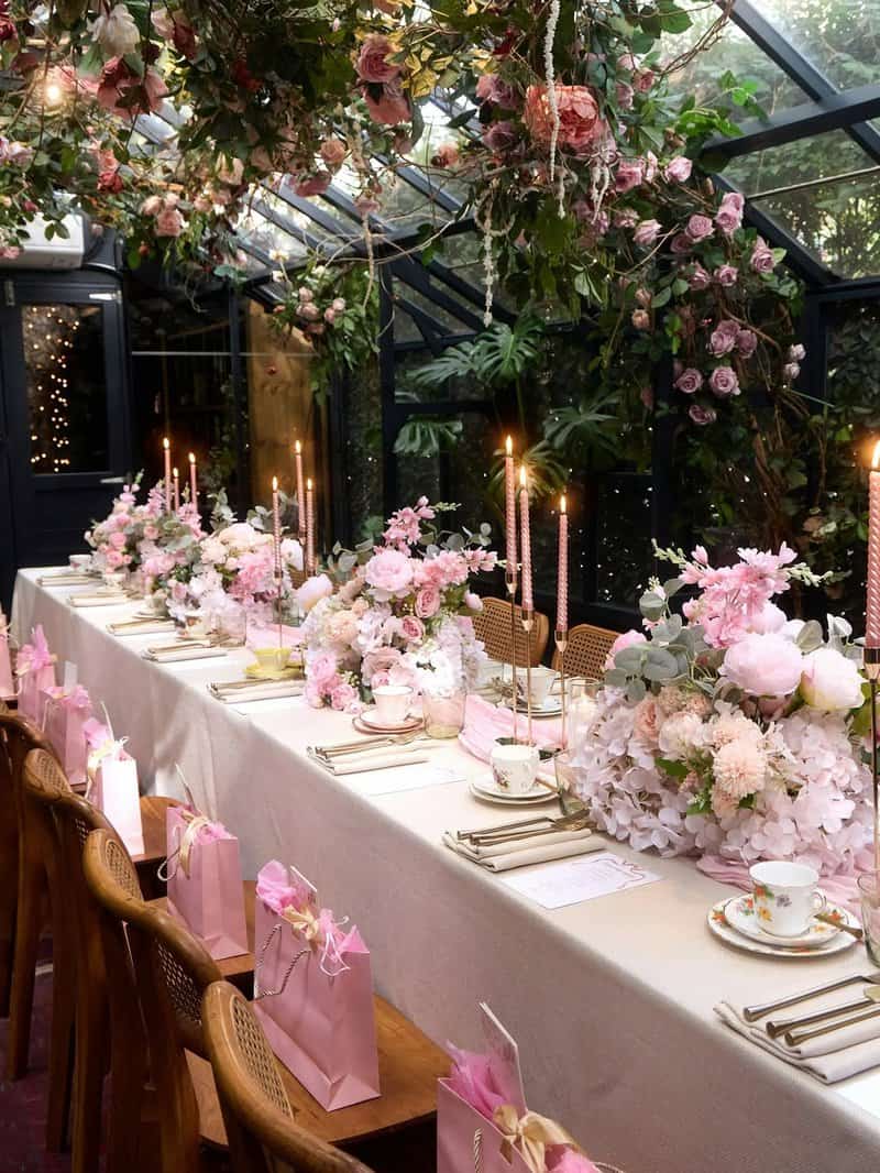 Long dining table inside a glasshouse with abundant pink floral arrangements, tall taper candles, beige table linen, gold flatware, teacups and saucers, and pink gift bags lined along wooden chairs