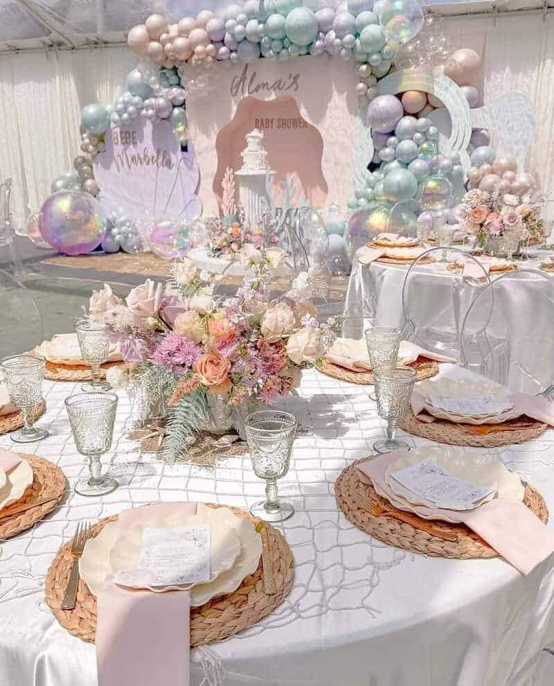 Round banquet tables with white linens, blush napkins, woven chargers, etched goblets, pastel floral centerpieces, and a large pastel balloon installation with iridescent spheres around a baby shower backdrop