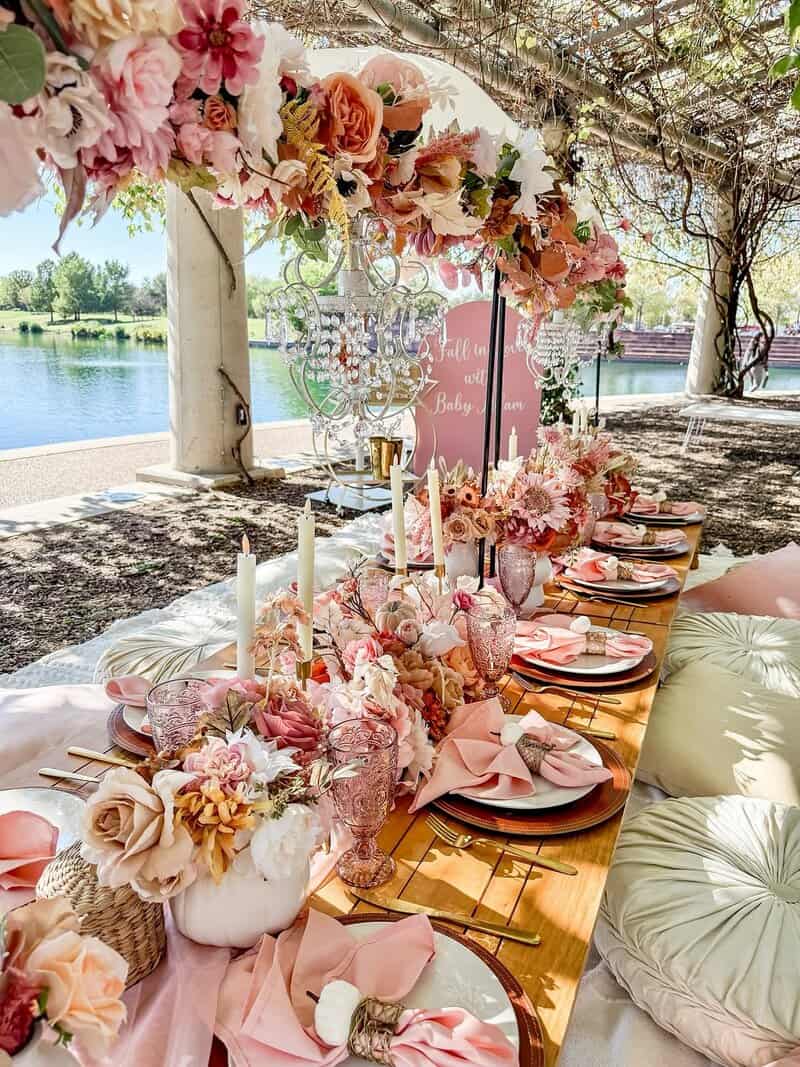 Outdoor picnic-style setup by a lake with a long low wooden table, pink and peach floral garlands overhead, crystal chandelier-style decor, pink glassware, candles, layered plates, floor cushions, and a pink welcome sign