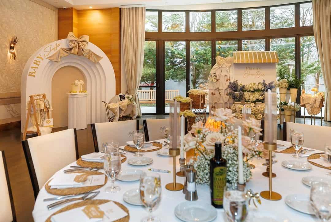 Indoor event space with round table set with gold-rimmed plates, gold candlesticks, glassware and woven chargers; in the background an arched baby shower backdrop with a bow and a flower bar display near large windows