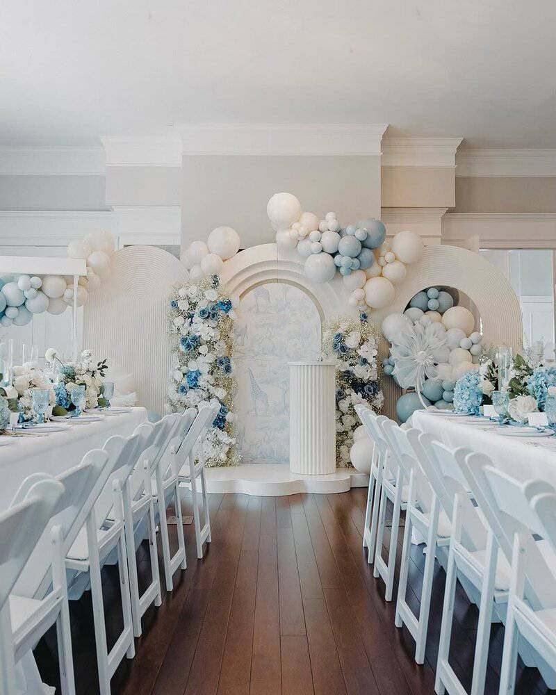 Bright indoor room with two long banquet tables and white folding chairs facing a central arched backdrop; pastel balloon garland in white and light blue surrounds floral arrangements and a column pedestal
