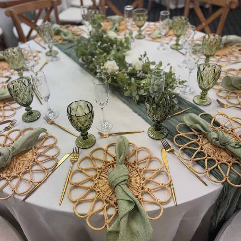 Round table with white tablecloth and sage-green gauze runner, rattan flower-petal chargers, sage knotted napkins, gold flatware, green goblets, champagne flutes, and a low greenery-and-white-flower centerpiece with wooden crossback chairs